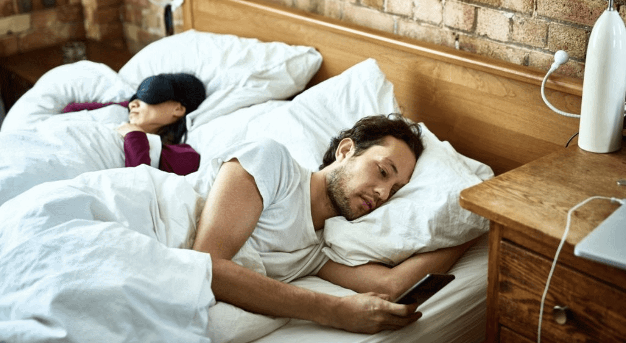 man looking at phone while partner sleeps with back turned to him